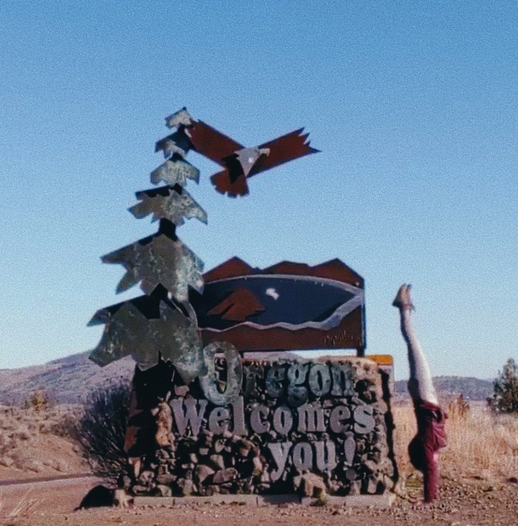 Jewel doing a handstand next to an "Oregon Welcomes You" sign and scultpure.